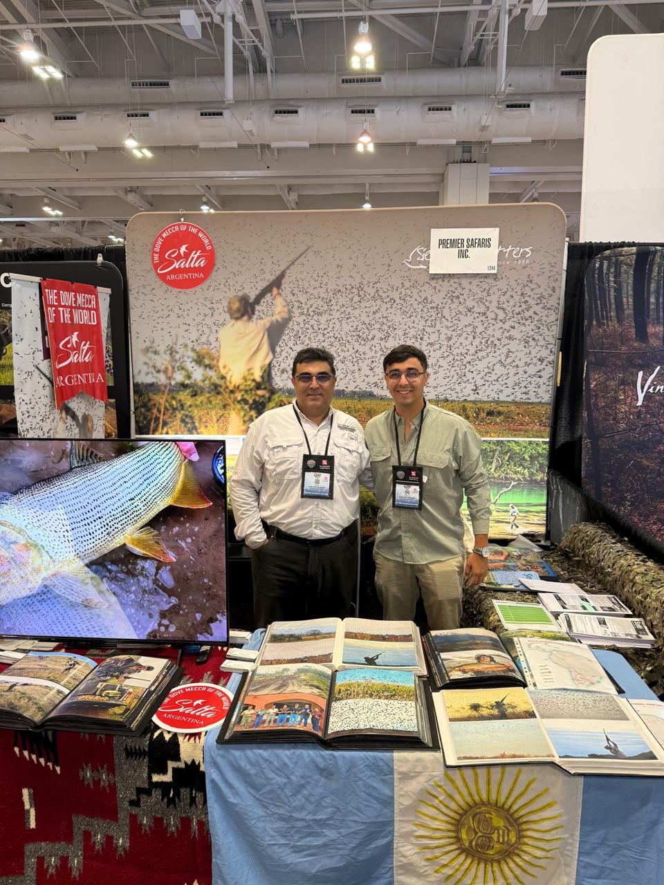 Dos personas junto a fotografías de caza y pesca en un stand de feria.